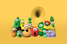 L’image montre un groupe de légumes et fruits animés, avec des visages souriants, comme dans un dessin animé. Ils sont alignés ensemble devant un grand fond jaune uni. Tous les personnages ont de grands yeux ronds, des bouches ouvertes en sourires, et ils semblent poser ensemble, comme pour une photo de groupe joyeuse. Derrière eux, on voit une grosse trompette ou un instrument à vent doré, comme si le groupe allait jouer de la musique.
